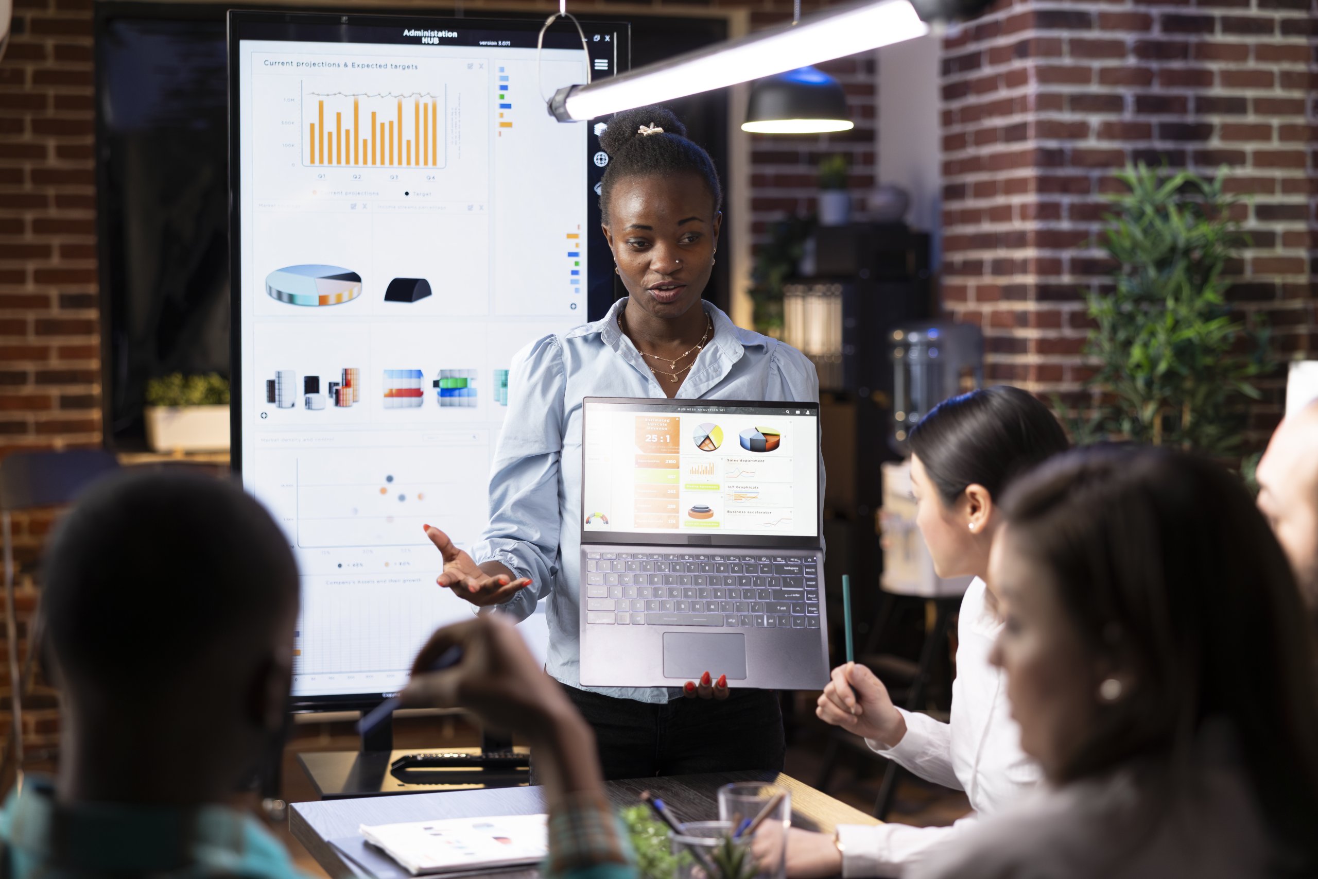 Startup team gathered around a table in a casual meeting room, discussing business strategy with laptops, charts, and digital tools in view.